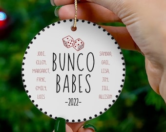 May include: White round Christmas ornament with black polka dots and a red dice design. The text "Bunco Babes -2022- " is printed in black. A list of names is printed in pink.