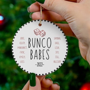 May include: White round Christmas ornament with black polka dots and a red dice design. The text "Bunco Babes -2022- " is printed in black. A list of names is printed in pink.