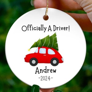 May include: A white ceramic Christmas ornament with a red car carrying a green Christmas tree on top. The ornament says "Officially A Driver!" with the name "Andrew" and the year "2024" below the car.