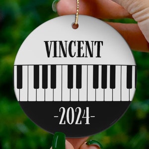 May include: A white and black round Christmas ornament with a piano keyboard design. The ornament has the name "VINCENT" and the year "2024" printed on it.