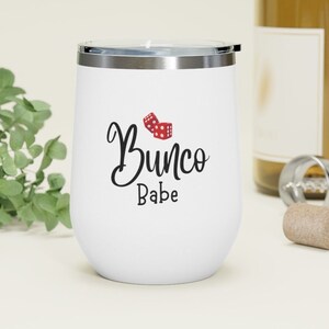Puede incluir: Un vaso de vino de acero inoxidable blanco con un dise&ntilde;o negro y rojo que dice "Bunco Babe" con dos dados rojos.