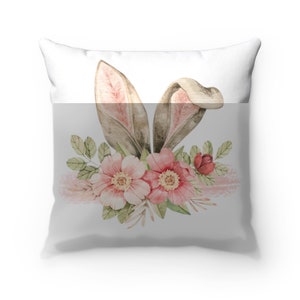 May include: A white pillow with a watercolor design of bunny ears and pink flowers. The bunny ears are brown with pink insides. The flowers are pink with green leaves.