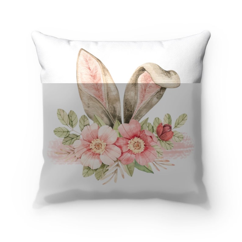 May include: A white pillow with a watercolor design of bunny ears and pink flowers. The bunny ears are brown with pink insides. The flowers are pink with green leaves.