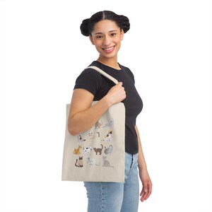 May include: A beige tote bag with a design of various cats in different poses. The bag has a natural cotton canvas texture and is being held by a person wearing a black t-shirt and blue jeans.