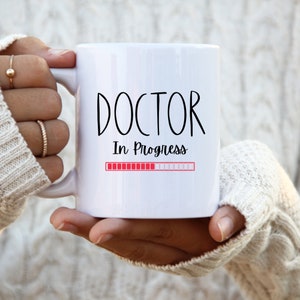 Könnte beinhalten: Weiße Keramiktasse mit schwarzem Text "DOCTOR In Progress" und einem roten Fortschrittsbalken.