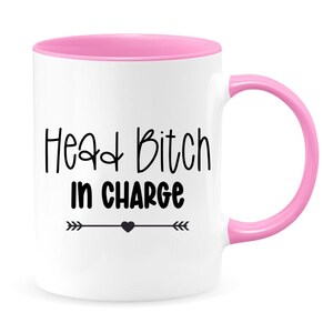 May include: White ceramic coffee mug with a pink rim and handle. The mug has black text that reads "Head Bitch in Charge" with a small heart and arrows on either side.