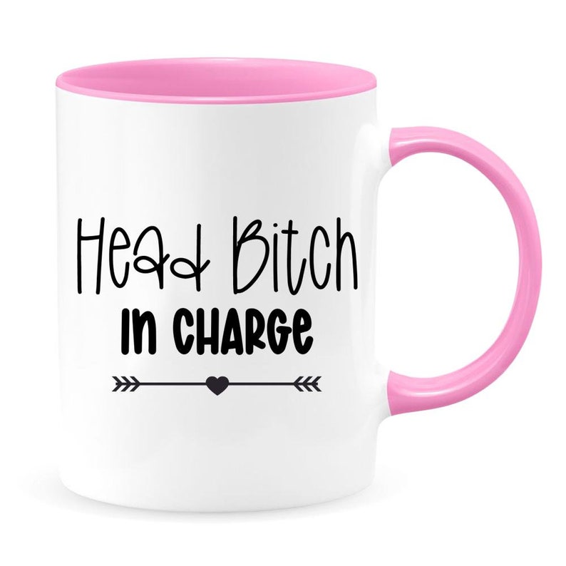 May include: White ceramic coffee mug with a pink rim and handle. The mug has black text that reads "Head Bitch in Charge" with a small heart and arrows on either side.