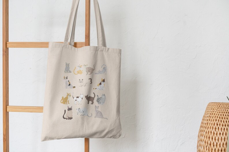 May include: A beige canvas tote bag with a design of various cats in different poses and colors. The bag has two long handles and is hanging on a wooden rack.