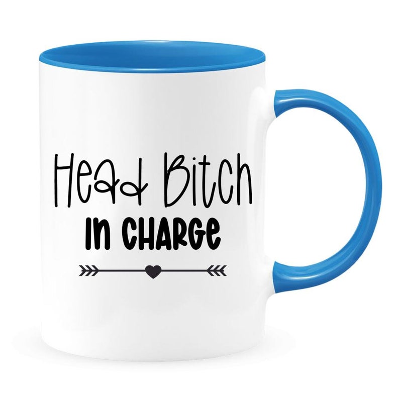 May include: White ceramic coffee mug with a blue rim and handle. The mug has black text that reads "Head Bitch in Charge" with a small black heart and arrows on either side.