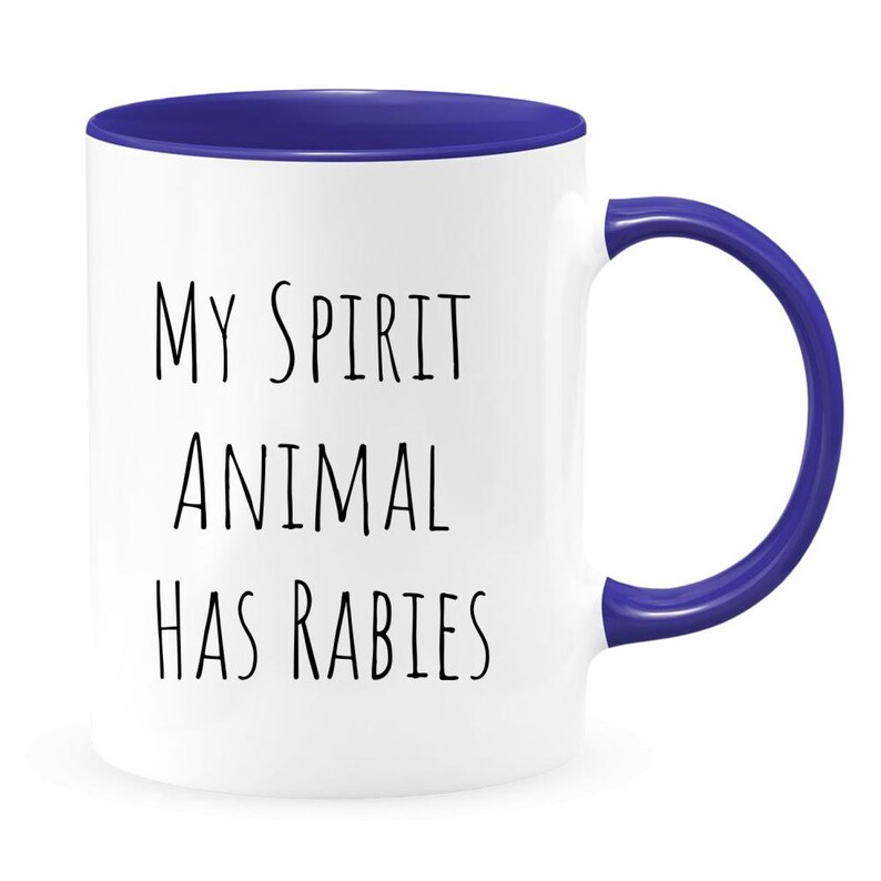May include: White ceramic coffee mug with a blue rim and handle. The mug has black text that reads "My spirit animal has rabies".