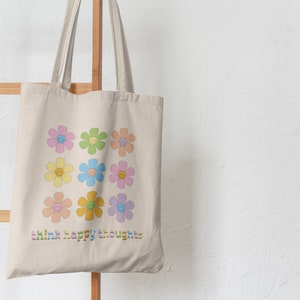 May include: A beige tote bag with a design of colorful flowers with smiley faces. The text "think happy thoughts" is printed on the bag.