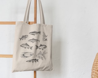 Puede incluir: Una bolsa de lona beige con ilustraciones en blanco y negro de varios peces. La bolsa tiene dos asas y está colgada de una escalera de madera.