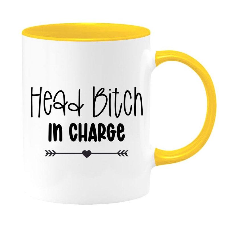 May include: White ceramic mug with a yellow rim and handle. The mug has black text that reads "Head Bitch in Charge" with a small black heart and arrows on either side.