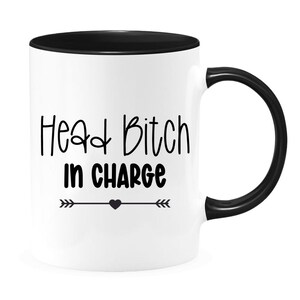 May include: White ceramic coffee mug with a black rim and handle. The mug has black text that reads "Head Bitch in Charge" with a small heart and arrows on either side.