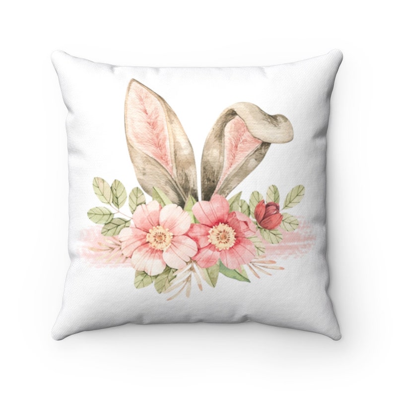 May include: A white throw pillow with a watercolor design of bunny ears and pink flowers. The bunny ears are brown with pink inside and the flowers are pink with green leaves.
