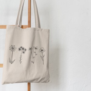 以下が含まれることがあります： 前面に黒い線の花の絵が描かれたベージュのキャンバス地のトートバッグ。バッグは木製ラックに掛けられています。
