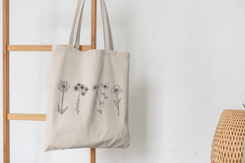 以下が含まれることがあります： 前面に黒い線の花の絵が描かれたベージュのキャンバス地のトートバッグ。バッグは木製ラックに掛けられています。
