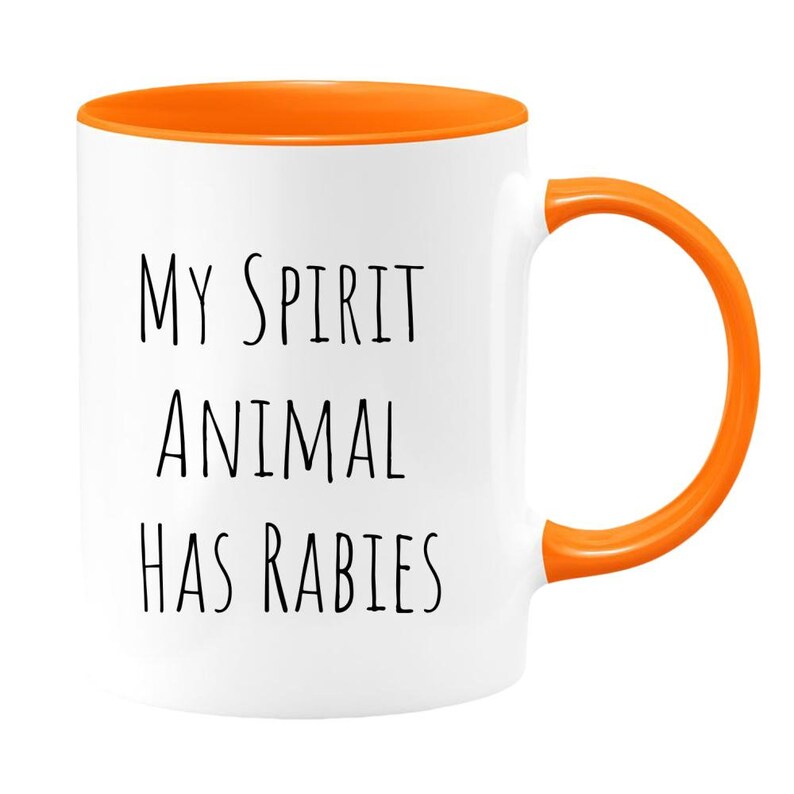 May include: White ceramic coffee mug with an orange rim and handle. The mug has black text that reads "MY SPIRIT ANIMAL HAS RABIES".
