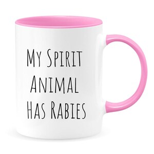 May include: White ceramic coffee mug with a pink rim and handle. The mug has black text that reads "My spirit animal has rabies".