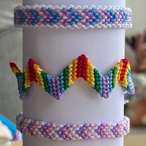 Op de afbeelding: Een verzameling kleurrijke handgemaakte vriendschapsarmbanden. De armbanden worden getoond op een witte cilinder, met verschillende patronen en kleurencombinaties, waaronder regenboog-, groen-wit- en rood-wit-ontwerpen.