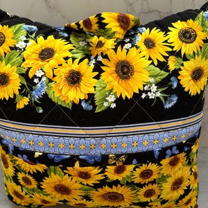 May include: A quilted tote bag featuring a vibrant sunflower pattern on a black background. The bag has a top handle and is decorated with yellow, green, and blue floral designs. The bag is approximately 14 inches wide and 12 inches tall.