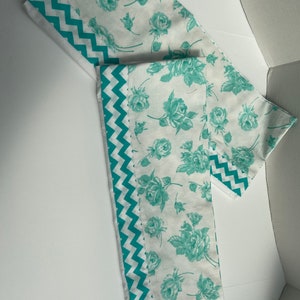 May include: Two white fabric panels with a teal floral pattern and a teal and white chevron border. The panels are folded in half and are lying on a white surface.