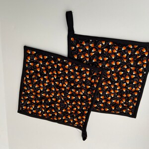 May include: Two black quilted pot holders with a Halloween candy corn pattern. The pot holders have a black border and a hanging loop.