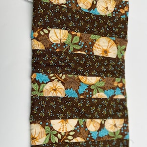 May include: A brown quilted fabric table runner with a pattern of white pumpkins, blue leaves, and brown branches. The runner has a quilted texture and is perfect for fall decor.