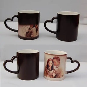 May include: Two black magic mugs with heart-shaped handles. One mug features a photo of a couple embracing, while the other mug is blank. The mugs are on a white surface.