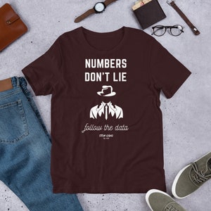 May include: A brown t-shirt with white text that reads "Numbers Don't Lie" and "follow the data". The text is in a stylized font and includes a graphic of a person wearing a fedora and suit.