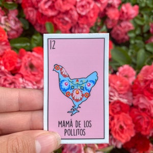 May include: A rectangular sticker featuring a blue chicken with floral patterns on a pink background. The number 12 is in the upper left corner, and the text "MAMÁ DE LOS POLLITOS" is at the bottom. The sticker is held in front of a backdrop of red roses.