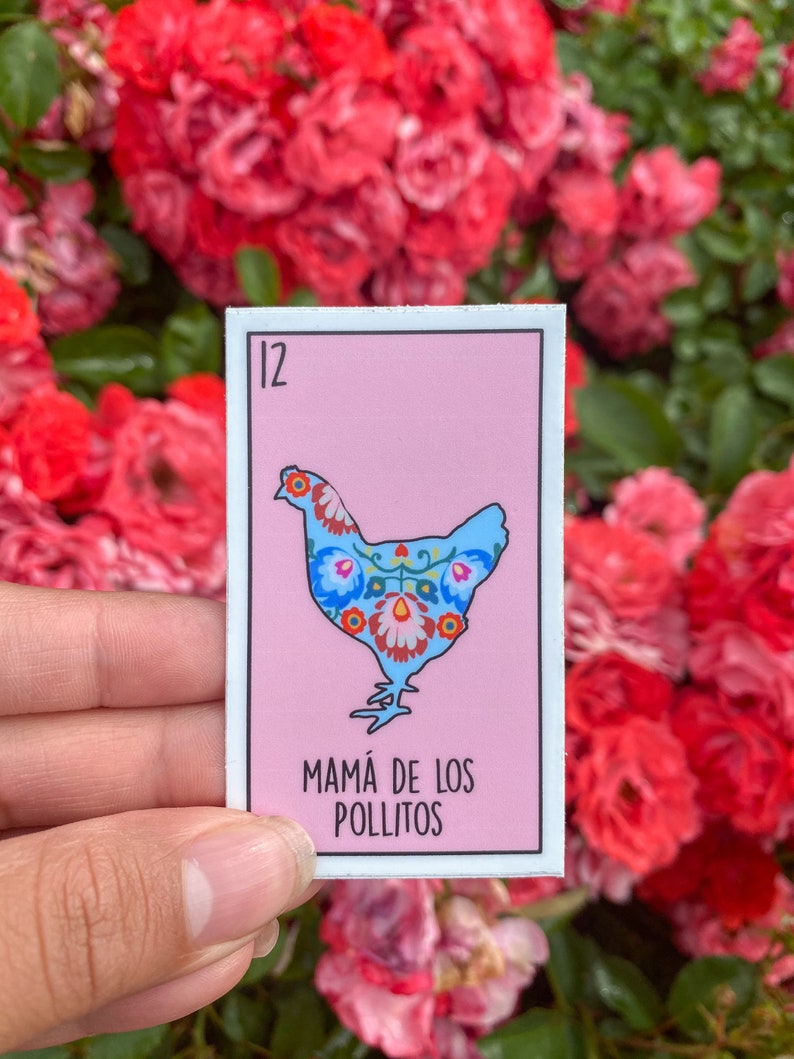 K&ouml;nnte beinhalten: Ein rechteckiger Aufkleber mit einem blauen H&uuml;hnerdesign und floralen Mustern auf rosa Hintergrund. Die Nummer 12 befindet sich in der oberen linken Ecke, und der Text "MAM&Aacute; DE LOS POLLITOS" steht unten. Der Aufkleber wird vor roten Rosen gehalten.