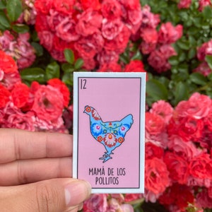 K&ouml;nnte beinhalten: Ein rechteckiger Aufkleber mit einer hellblauen H&uuml;hnerillustration und floralen Mustern auf rosa Hintergrund. Die Nummer 12 befindet sich oben links, und der Text "MAM&Aacute; DE LOS POLLITOS" steht darunter. Der Aufkleber wird vor roten Rosen gehalten.