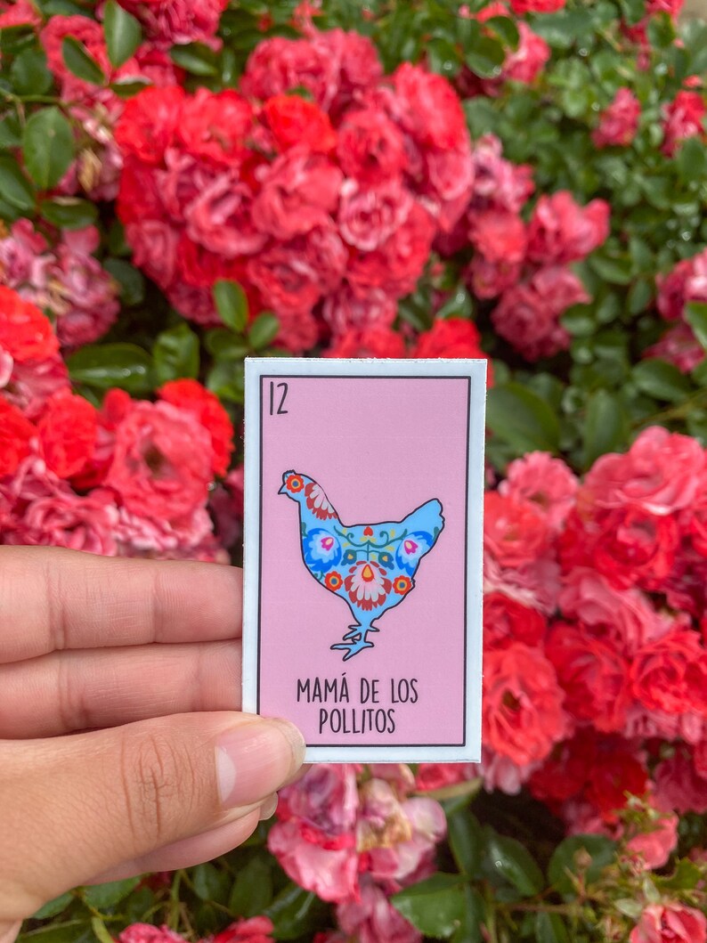 K&ouml;nnte beinhalten: Ein rechteckiger Aufkleber mit einer hellblauen H&uuml;hnerillustration und floralen Mustern auf rosa Hintergrund. Die Nummer 12 befindet sich oben links, und der Text "MAM&Aacute; DE LOS POLLITOS" steht darunter. Der Aufkleber wird vor roten Rosen gehalten.