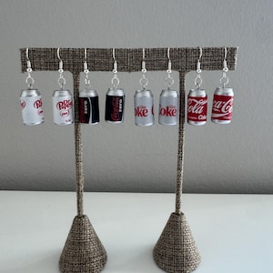 May include: Eight miniature soda can earrings hanging from silver-toned hooks. The collection includes Dr. Pepper, Diet Dr. Pepper, Coca-Cola Zero, Diet Coke, and classic Coca-Cola designs in red, white, black, and silver colors with brand logos.