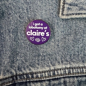 May include: A circular purple pin with white text that reads i got a lobotomy at claire's, accompanied by two white heart outlines and a small white brain icon in the center, attached to a blue denim fabric surface with visible stitching.