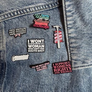 May include: A collection of seven enamel pins attached to blue denim fabric. The pins feature various slogans including "Strong women don't have attitudes they have standards," "very demure very mindful very softgirl," and "I support womens rights and wrongs."