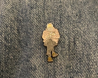May include: A silver-colored enamel pin depicting a seated figure wearing a coat, glasses, and a face mask, holding a pair of mittens. The pin is set against a blue denim background.