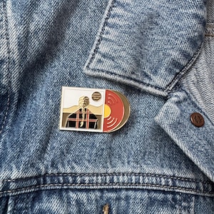 Op de afbeelding: Een rechthoekige emaille pin met een gestileerde afbeelding van een persoon en een vinylplaat. De pin heeft een gouden rand en is geplaatst op een spijkerjasje. De plaat is rood en goud.