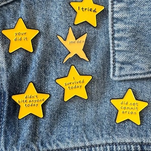 Puede incluir: Seis pines de esmalte amarillos en forma de estrella con texto en negro. Los pines incluyen las frases: I tried, your did it, your did it, I survived today, didn't bite anyone today y did not commit arson. Los pines están sujetos a tela vaquera azul.