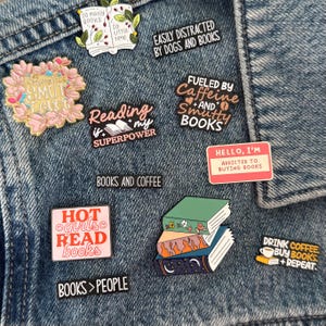 May include: A collection of enamel pins featuring book-themed designs and text including "So many books so little time," "Easily distracted by dogs and books," "Reading is my superpower," "Fueled by caffeine and smutty books," and "Hot girls read books."