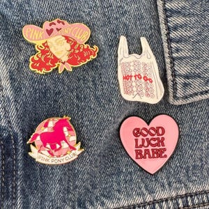 May include: Four enamel pins: one featuring a person in a pink cowboy hat with the text PINK PONY CLUB, one white plastic bag shape with the text HOT TO GO, one pink horse on a disco ball with PINK PONY CLUB, and one pink heart with GOOD LUCK BABE.