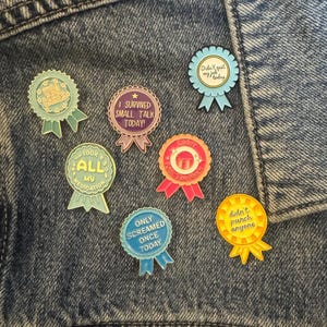 Könnte beinhalten: Eine Sammlung bunter Emaille-Anstecknadeln mit humorvollen Sprüchen auf einer Jeansjacke. Die Anstecknadeln haben die Form von Schleifen und enthalten Texte wie "Didn't quit my job today" und "Took all my medication."