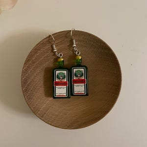 May include: A pair of novelty earrings shaped like miniature green and black liquor bottles. The earrings feature silver-colored fish hook ear wires and are displayed on a wooden plate. The bottles have a label with the word "Jägermeister".