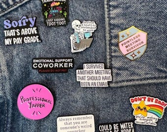 May include: A collection of enamel pins featuring humorous text and graphics, including a train, a skeleton at a computer, a shield, a pink circle, and a star with a rainbow, all pinned to a blue denim fabric surface.