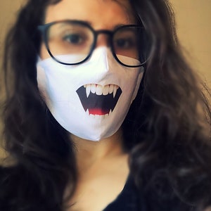 Puede incluir: Una mascarilla facial blanca con un diseño de dientes de vampiro negros y rojos. La mascarilla la lleva puesta una persona con el pelo largo y oscuro y gafas con montura negra.