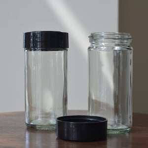 Op de afbeelding: Twee glazen potten met zwarte deksels. De potten zijn leeg en staan op een houten tafel.