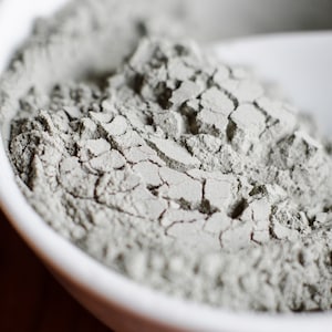 May include: A close-up of a white bowl filled with a light gray powder. The powder has a slightly textured surface and appears to be dry.