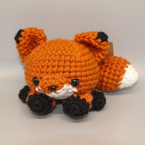 May include: A crocheted orange fox plush toy with black and white accents. The fox has a white belly and a white tip on its tail.