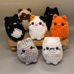 May include: A collection of seven crocheted cat figurines in various colors, including black, white, orange, and gray. Each cat has black eyes, a pink nose, and a smiling or slightly grumpy expression. The cats are displayed on a small wooden platform.